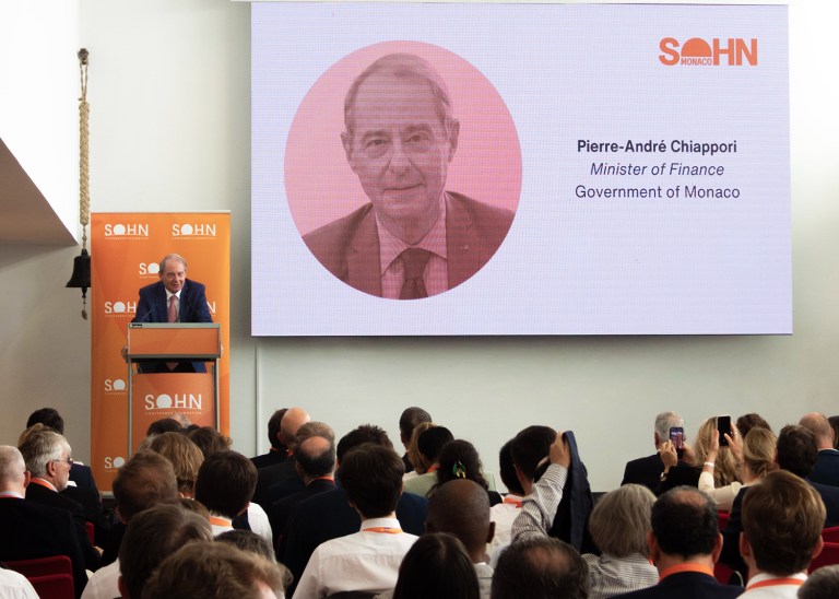 Crowded conference room at the Monaco Yacht Club, attendees listen to the Minister of Finance