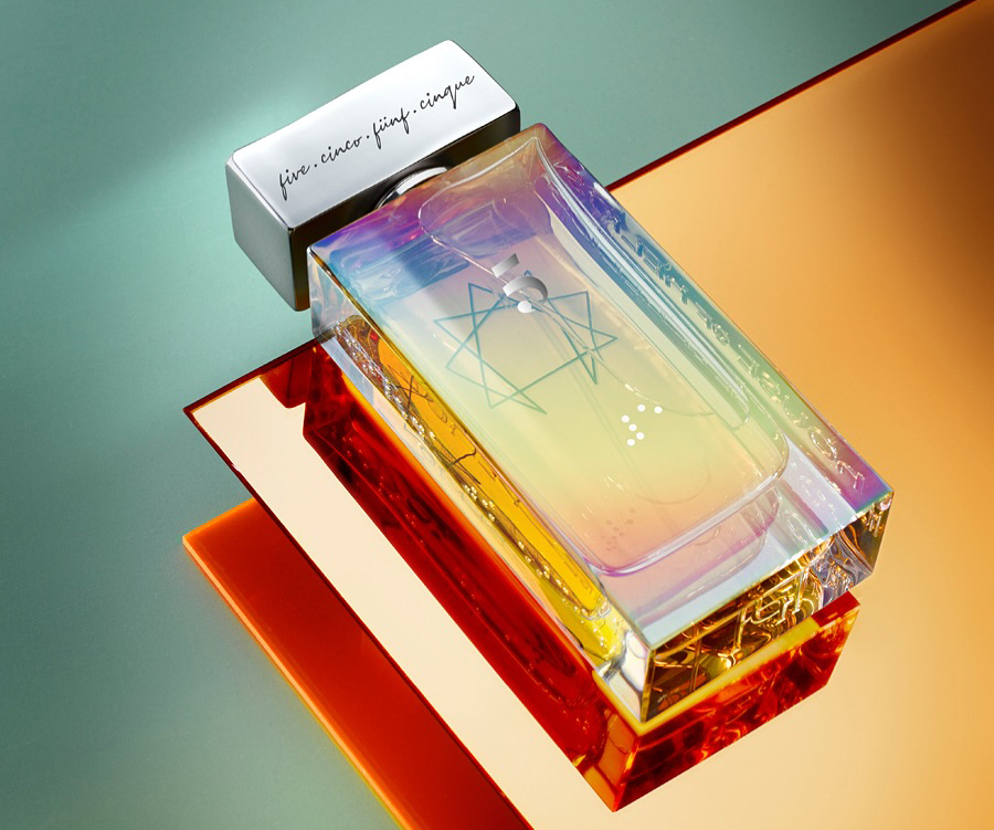 A prism-like perfume bottle with a rainbow gradient sits on reflective orange surfaces, evoking elegance and modernity. The cap is metallic with cursive script.