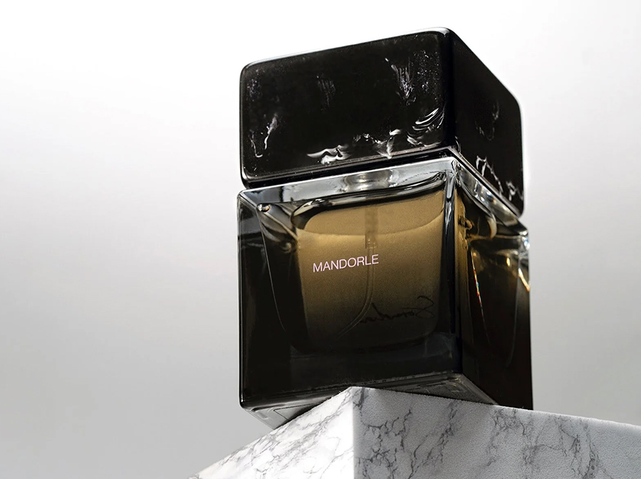 Elegant perfume bottle labeled "Mandorle" with a sleek black cap, placed on a marble pedestal. Sophisticated and luxurious ambiance.