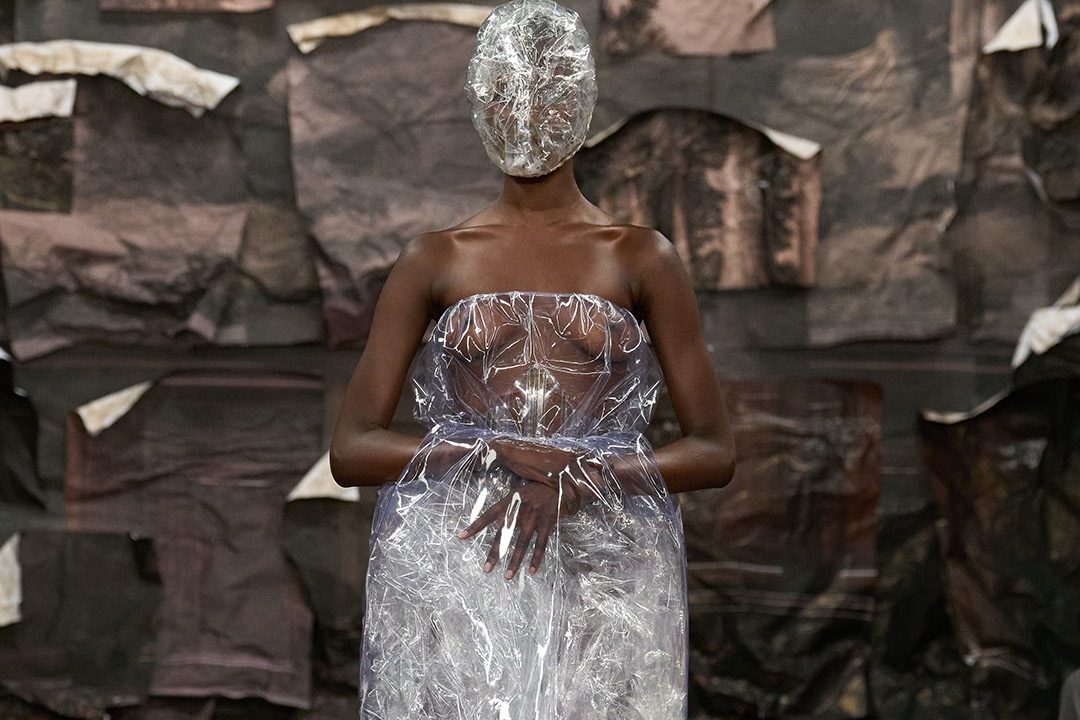 Black runway model wears a dress made of transparent clear plastic. Her face is cover with a plastic mask.