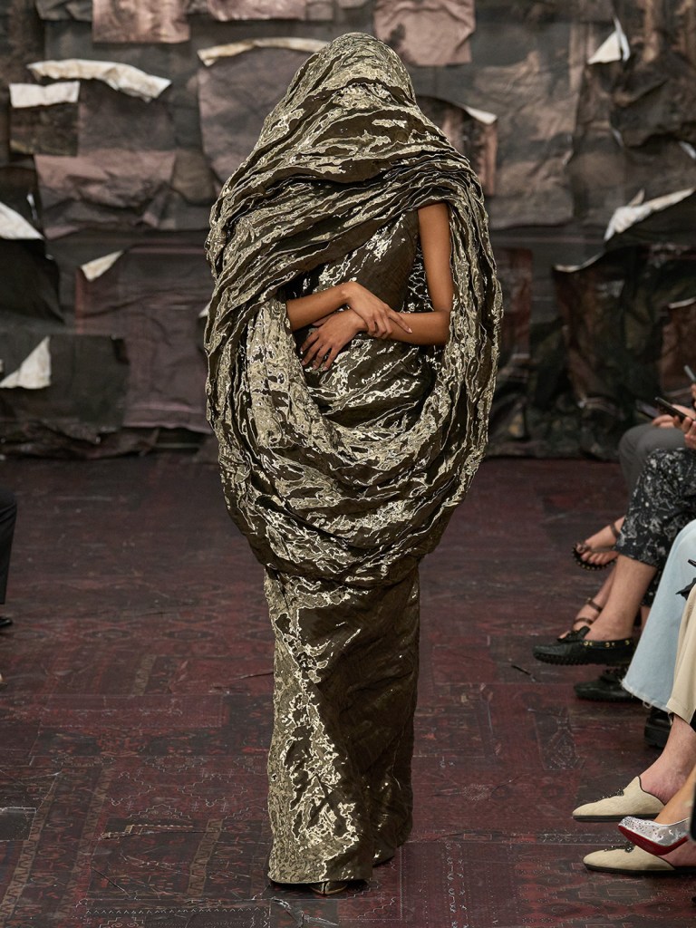 model on the runway wearing metallic gown, her face is covered with the same fabric.