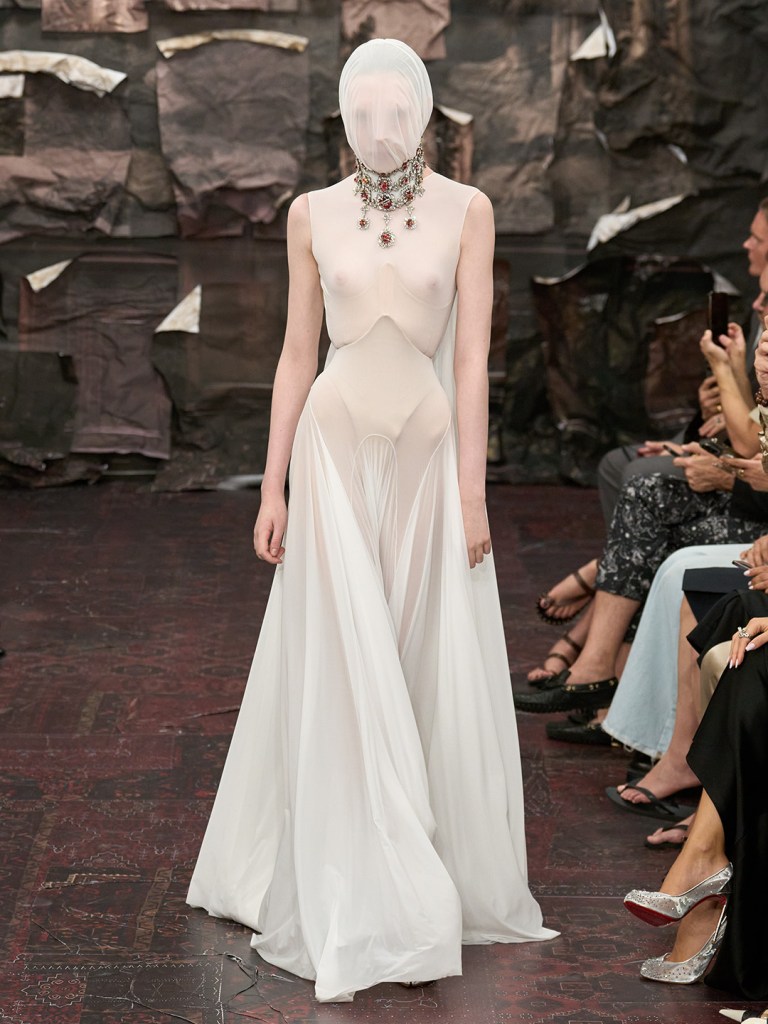 catwalk model is walking the runway wearing sheer milky coloured dress with large stone jewellery