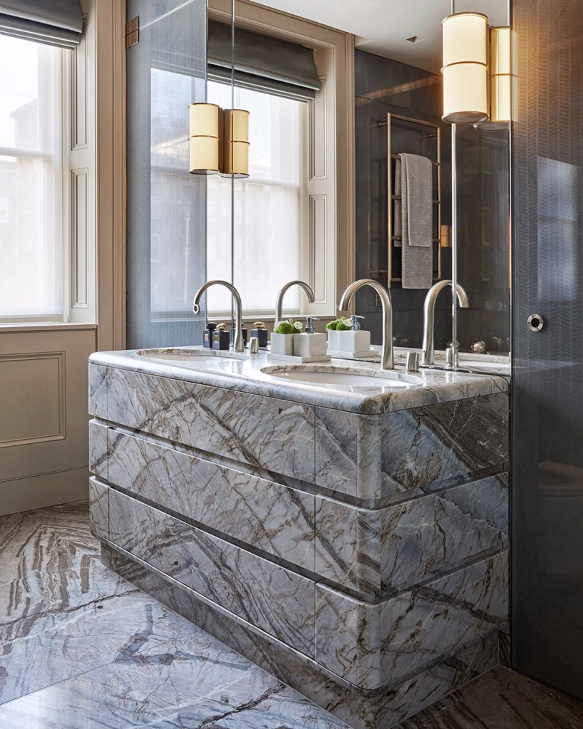 Chic interior design of a bathroom featuring grey marble flooring sink tub, silver taps, mirror, golden lamps and a window.