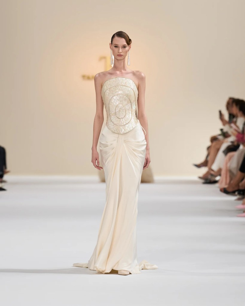 A model walks down the runway showcasing an elegant cream gown with a structured bodice and draped silk skirt.