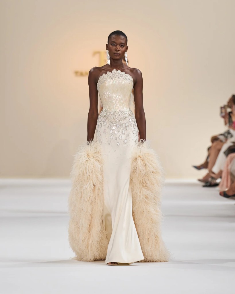 A model walks the runway wearing a strapless cream gown adorned with intricate beading and mother-of-pearl details, complemented by a dramatic ostrich feather shrug.
