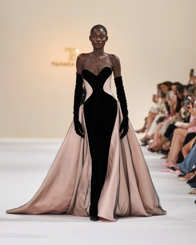 A model walks the runway showcasing a dramatic black and blush evening gown with long gloves.