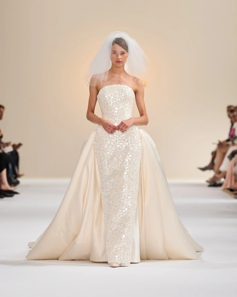 Model showcasing a cream-colored wedding gown with a form-fitting bodice and a flowing skirt, complemented by a delicate veil, walking down the runway.
