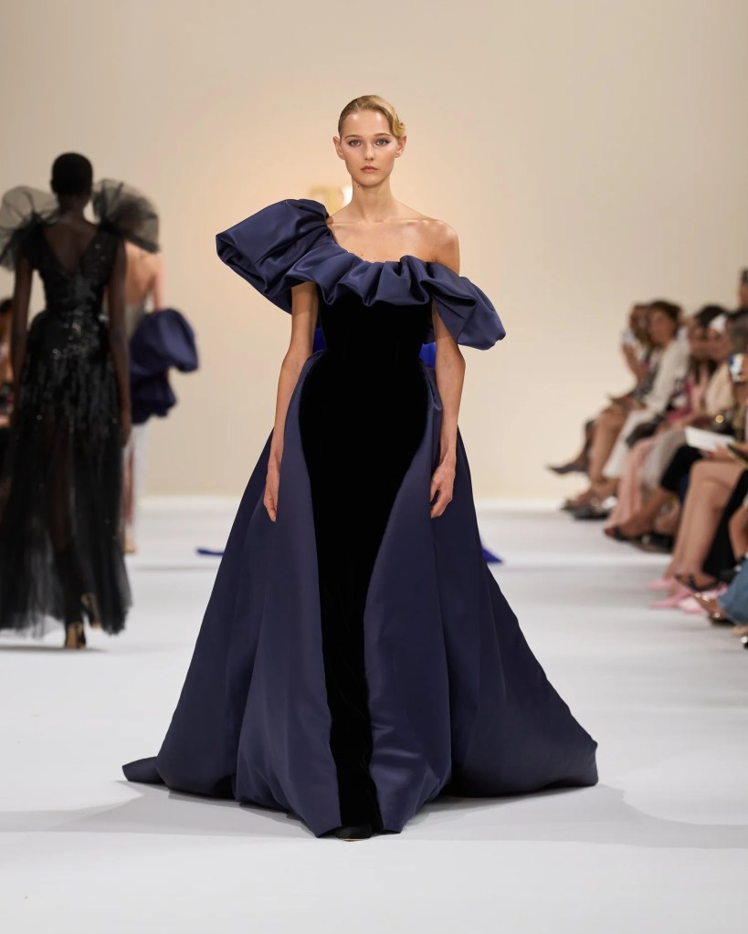 A model showcases an elegant navy gown with a dramatic ruffled neckline, walking down the runway in a fashion show.