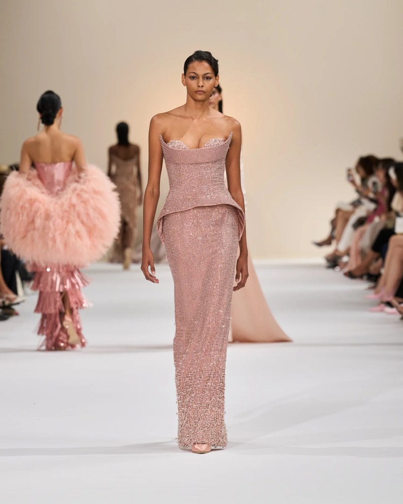 A model walks the runway in a stunning blush pink gown with intricate beading and a structured silhouette.