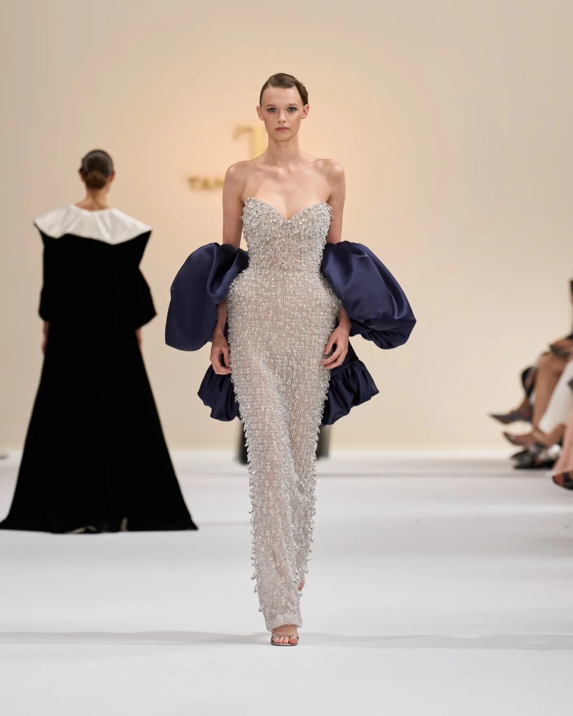 A model walks the runway in a shimmering silver gown adorned with intricate beading and navy puff sleeves.