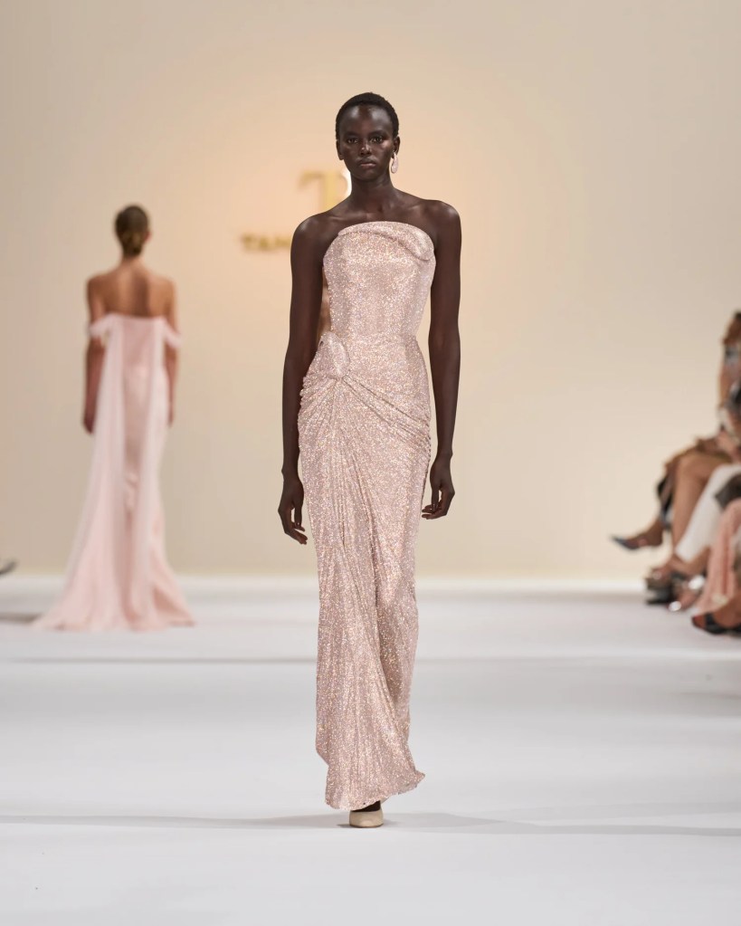 Model walking down the runway in a shimmering blush gown with a fitted silhouette and draped detailing.