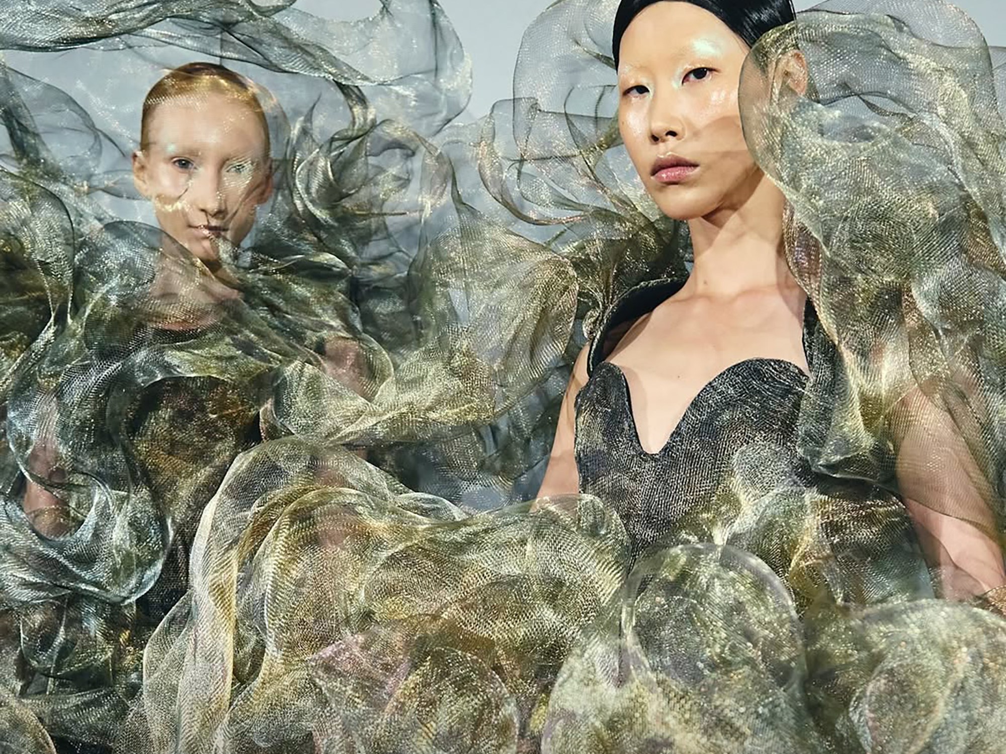 Two models in extravagantly flowing, translucent dresses with wave-like patterns and metallic hues stand with calm, confident expressions. Fashion show ambiance