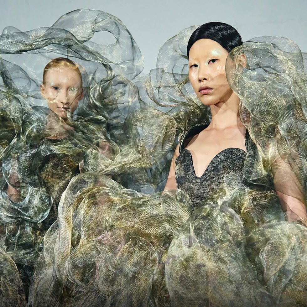 Two models in extravagantly flowing, translucent dresses with wave-like patterns and metallic hues stand with calm, confident expressions. Fashion show ambiance