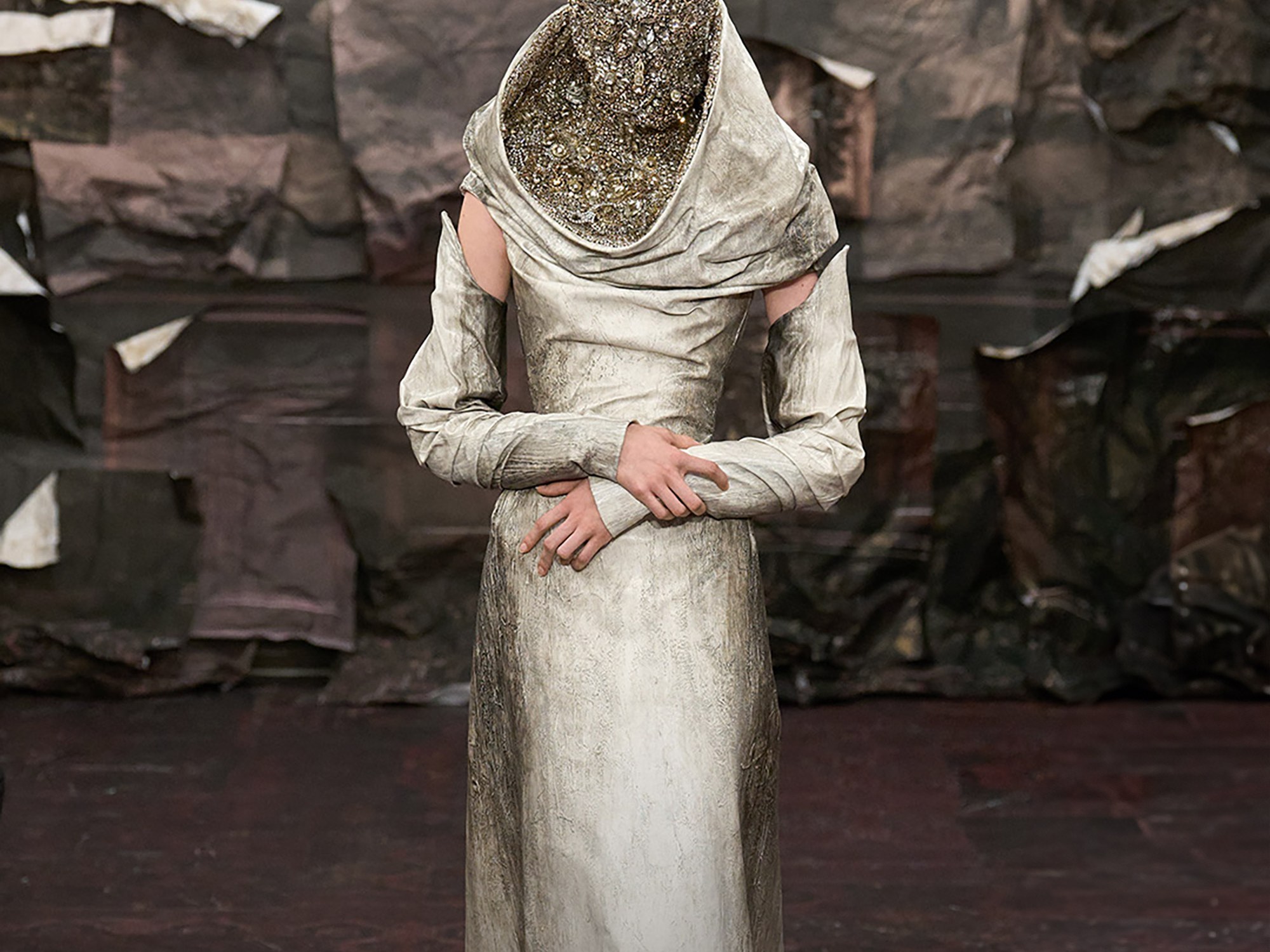 fashion model standing still on the catwalk with her hands crossed, wearing metallic coloured beige dress and face mask embellished with swarovski stones