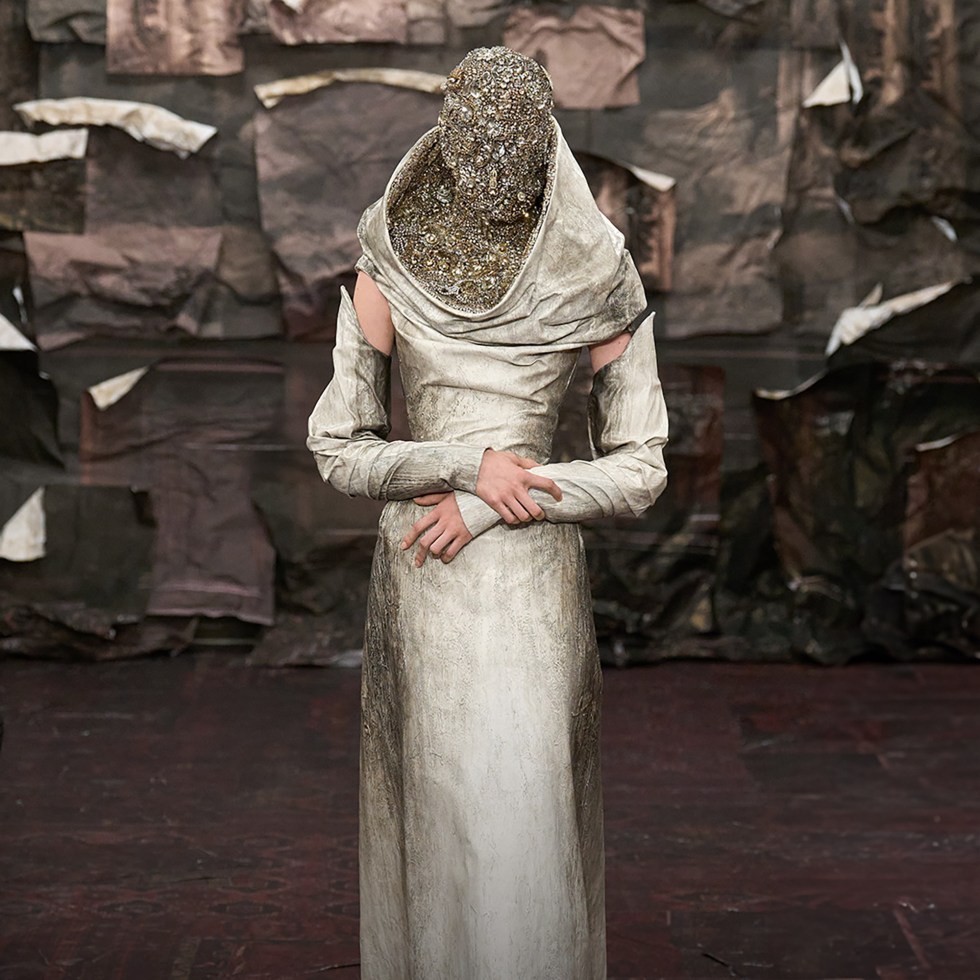 fashion model standing still on the catwalk with her hands crossed, wearing metallic coloured beige dress and face mask embellished with swarovski stones