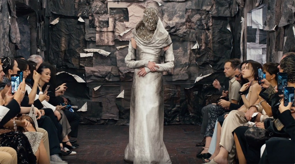 front row guests viewing and filming with their mobile phones a catwalk model wearing a metallic silk gown and embellished face and head cover in rhinstones
