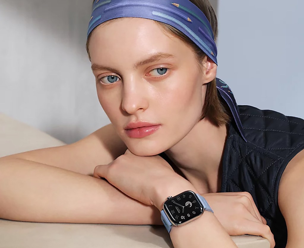 A close-up of a woman wearing a stylish Apple Watch Hermès on her wrist, with a light blue watch strap and a patterned silk headband, showcasing a modern aesthetic.