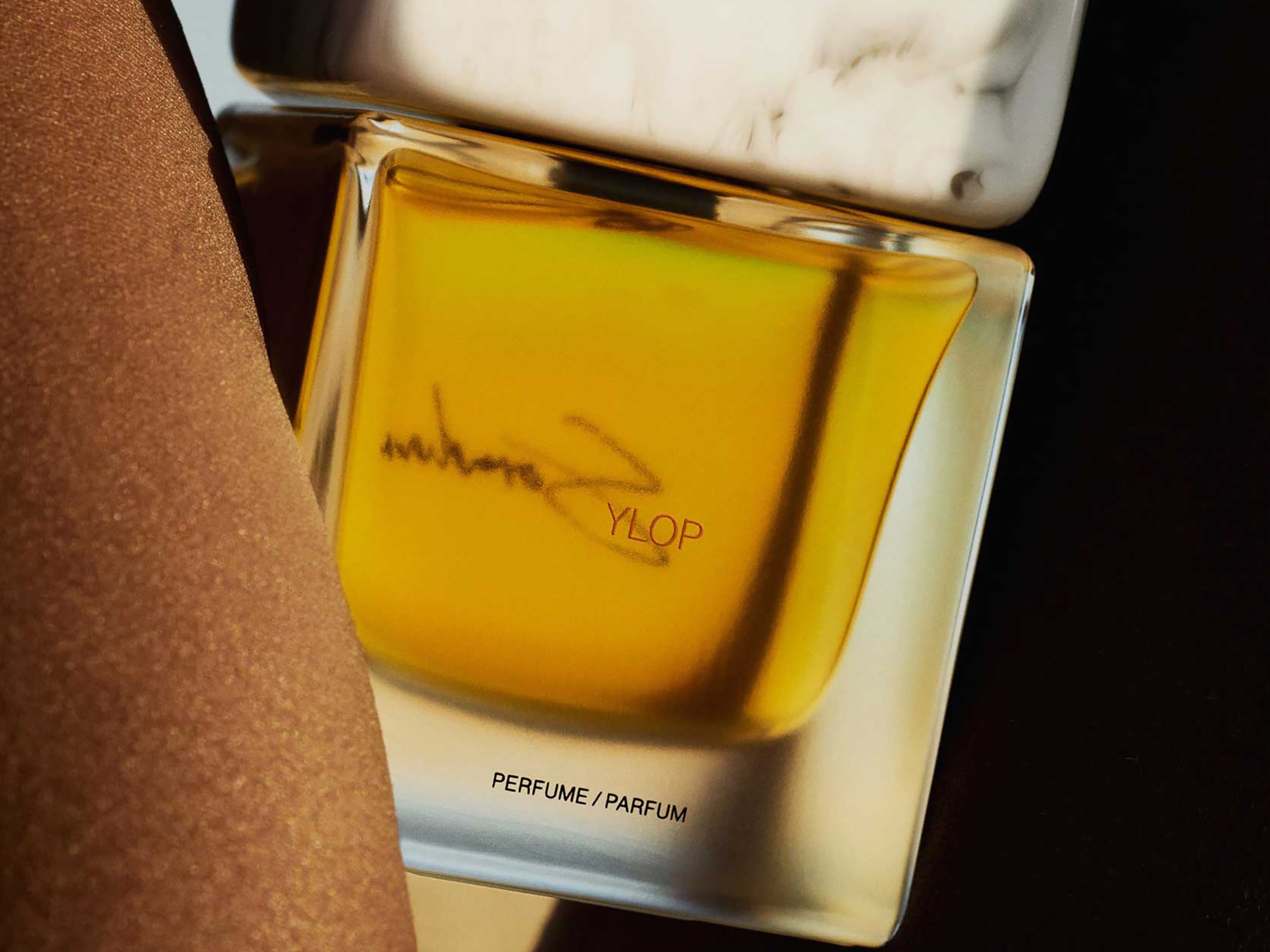 A golden-yellow perfume bottle with a marbled white cap is angled between smooth, brown surfaces. The mood is luxurious and sophisticated.