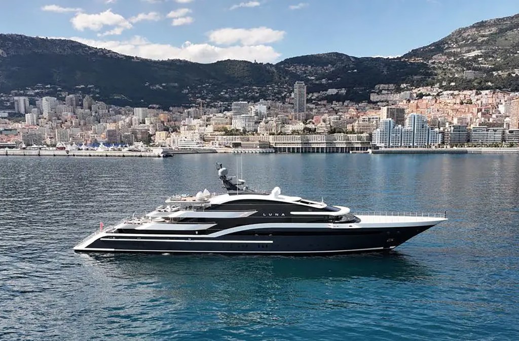 A luxurious yacht named Luna glides on a clear blue sea with a vibrant cityscape and mountainous backdrop. The scene is serene and opulent.