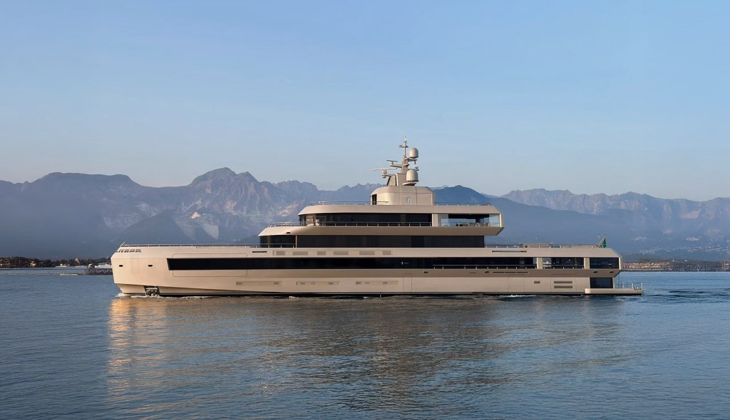 A luxurious golden yacht sails on calm waters with majestic mountains in the background under a clear blue sky, conveying elegance and tranquility.