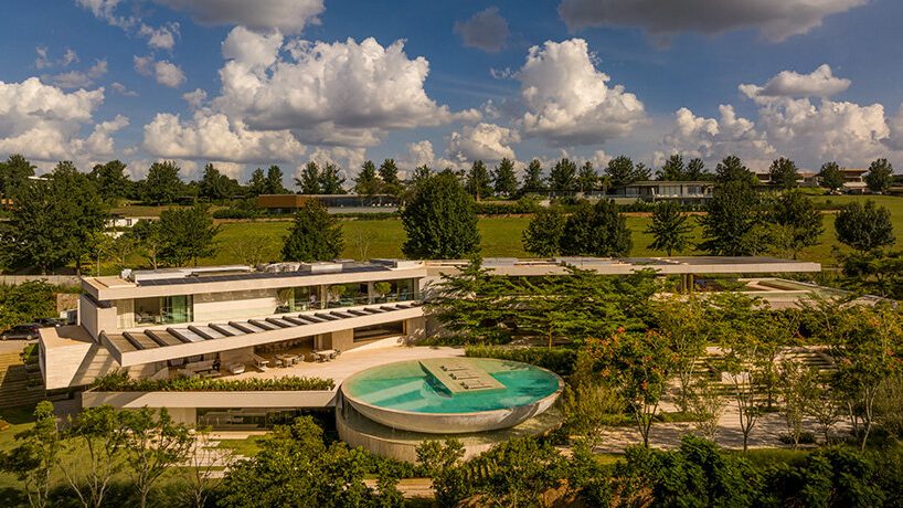 Modern mansion with a circular pool surrounded by lush greenery under a vibrant blue sky with fluffy clouds, conveying a serene and luxurious ambiance.