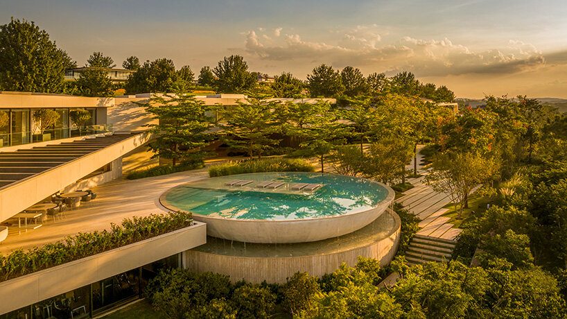 A modern building with a circular rooftop pool surrounded by lush greenery. Sunlit, with a serene ambiance and a backdrop of trees and a cloudy sky.