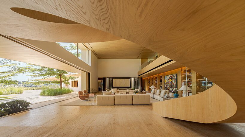 Spacious modern interior with wooden ceiling and walls, large windows, and beige sofas. Open design connects to an outdoor view, creating a tranquil and airy atmosphere.