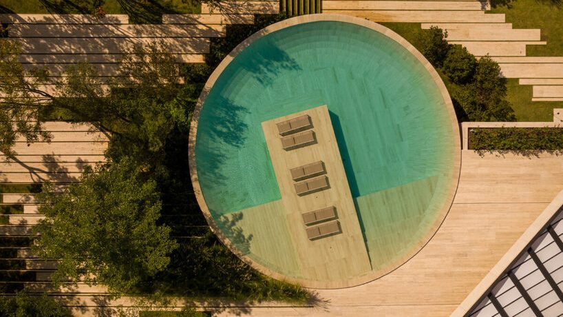 Aerial view of a circular pool with a central wooden platform holding three lounge chairs. Surrounding greenery and paved pathways add serenity and texture.