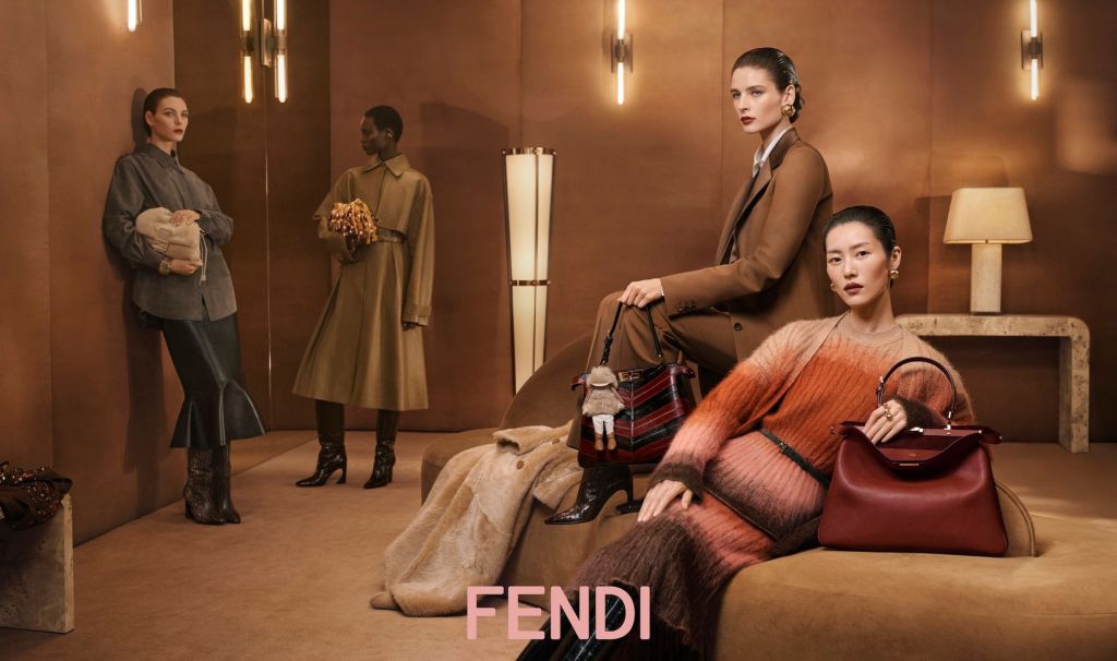 Elegant fashion scene in a modern room with warm brown tones. Four women in stylish outfits pose with luxury handbags, exuding sophistication. Fendi logo displayed.
