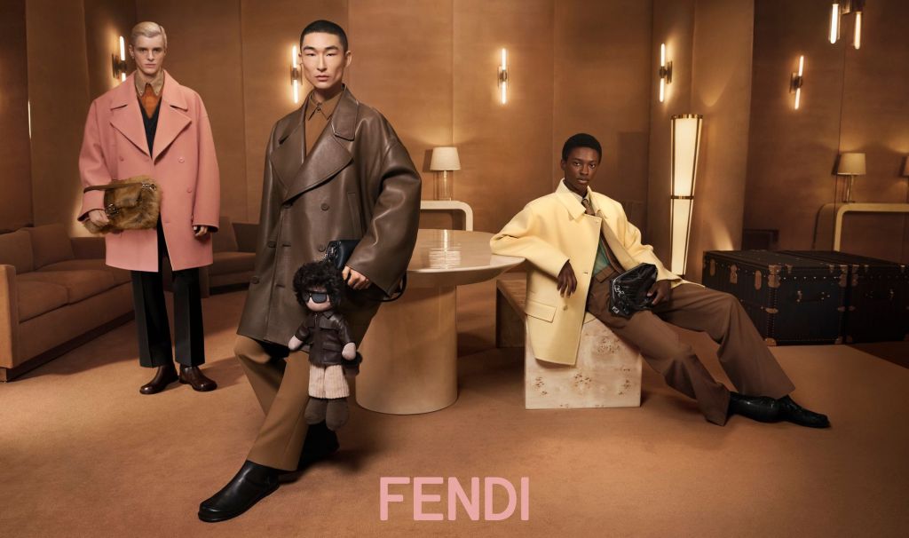 Three models in a luxurious room wearing Fendi outfits. One in pink, another in brown with a puppet, and the third in yellow. Elegant ambiance.