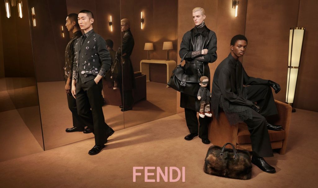 Three models pose in a luxurious, warmly lit room with brown tones. One stands against a mirror, one holds a designer bag, and another sits on an armchair. Fendi logo is at the bottom.
