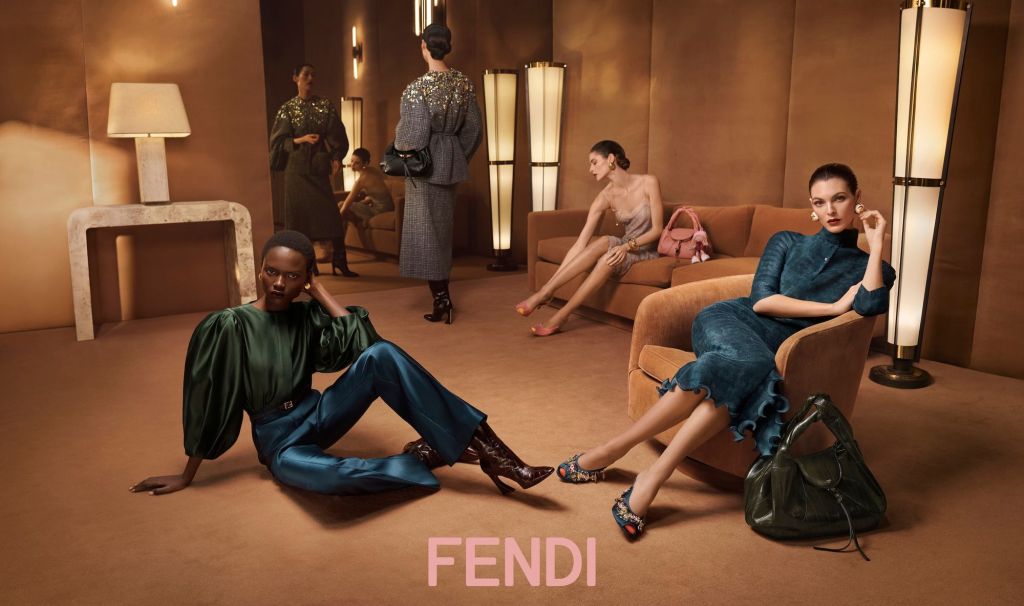 Luxurious fashion scene with four models in elegant outfits. A chic room with warm lighting, artful poses, and a sophisticated vibe. Fendi branding visible.