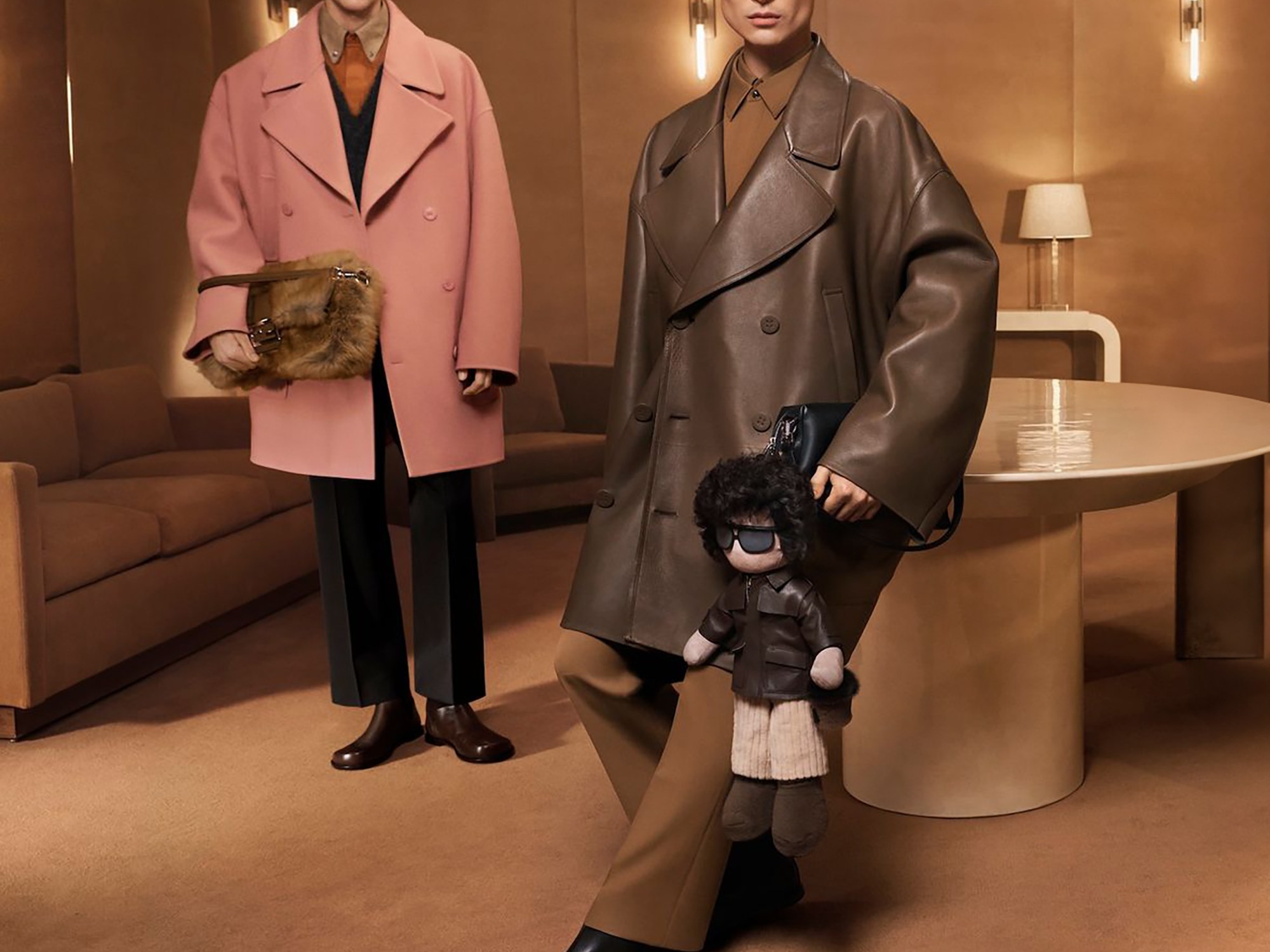 Two models pose in a stylish room; one in a pink coat and fur bag, the other in a brown leather coat holding a puppet. Elegant and modern setting.