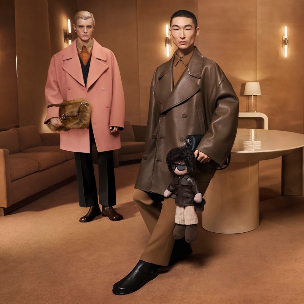 Two models pose in a stylish room; one in a pink coat and fur bag, the other in a brown leather coat holding a puppet. Elegant and modern setting.