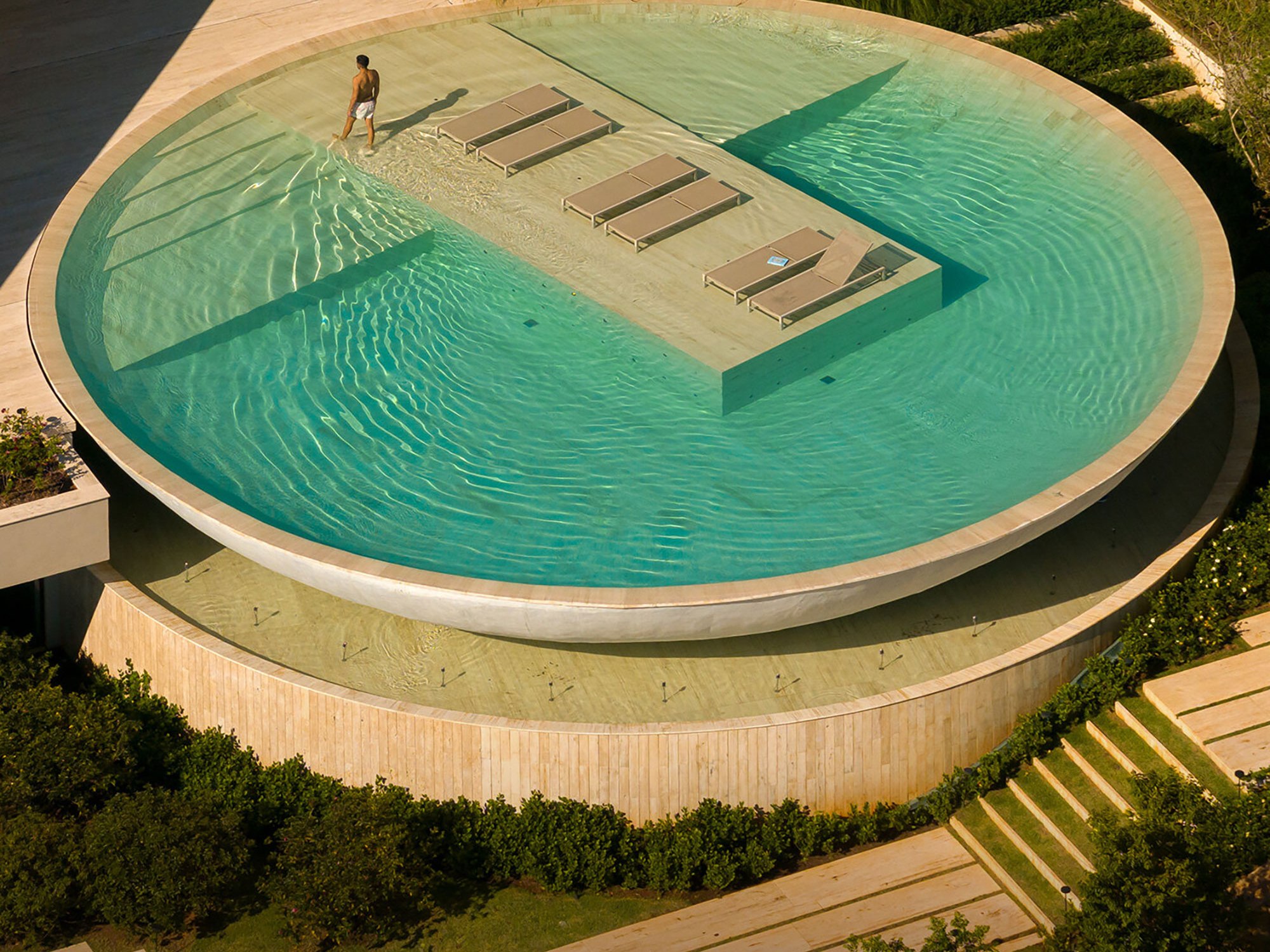 Aerial view of a large, round water feature with a modern design. A man walks on a pathway surrounded by water, with loungers in the center, evoking calm serenity.