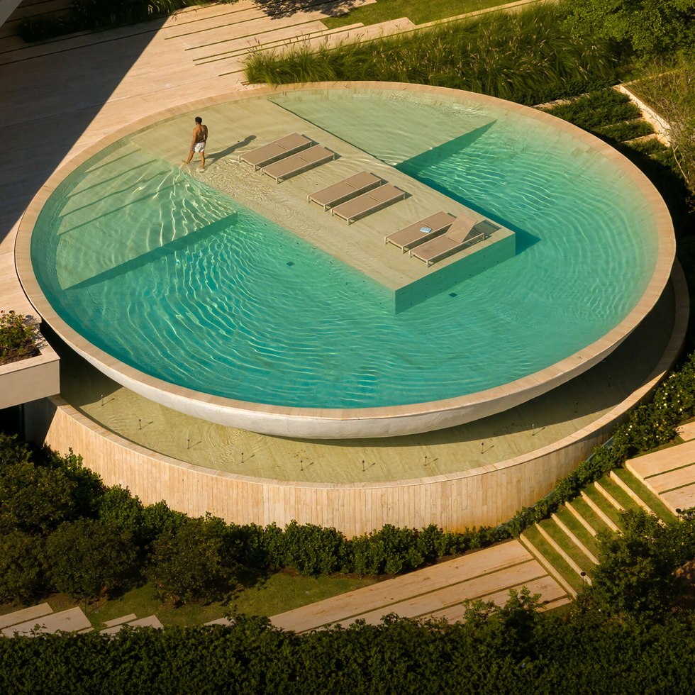 Aerial view of a large, round water feature with a modern design. A man walks on a pathway surrounded by water, with loungers in the center, evoking calm serenity.