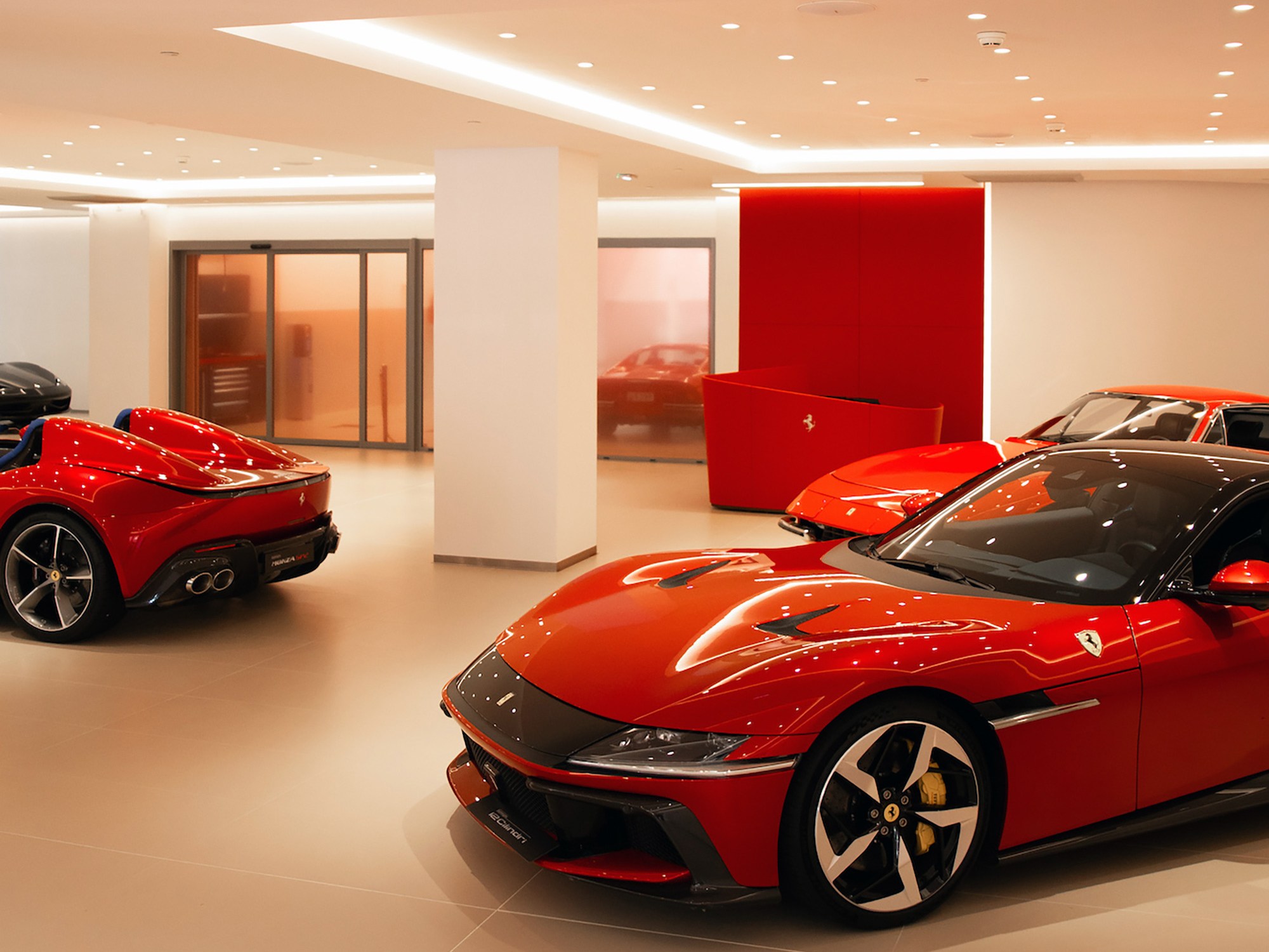 A showroom displaying sleek red sports cars, including convertibles, with glossy finishes under bright ceiling lights. The setting is elegant and luxurious.