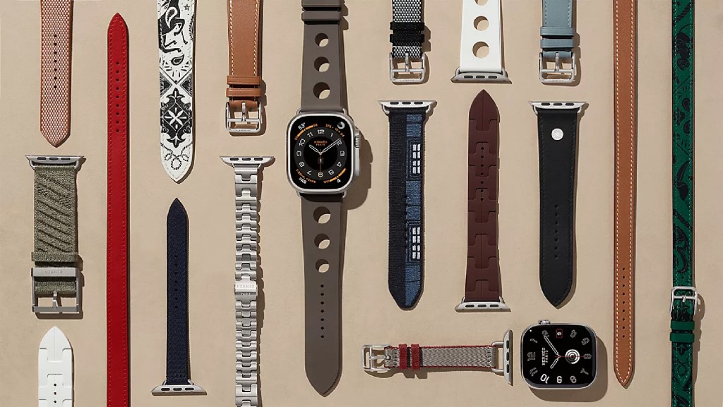 A flat lay image showcasing various Apple Watch bands and a smartwatch displaying a dark watch face. The bands come in different materials and colors, including leather, woven fabric, and silicone, arranged artistically on a neutral background.