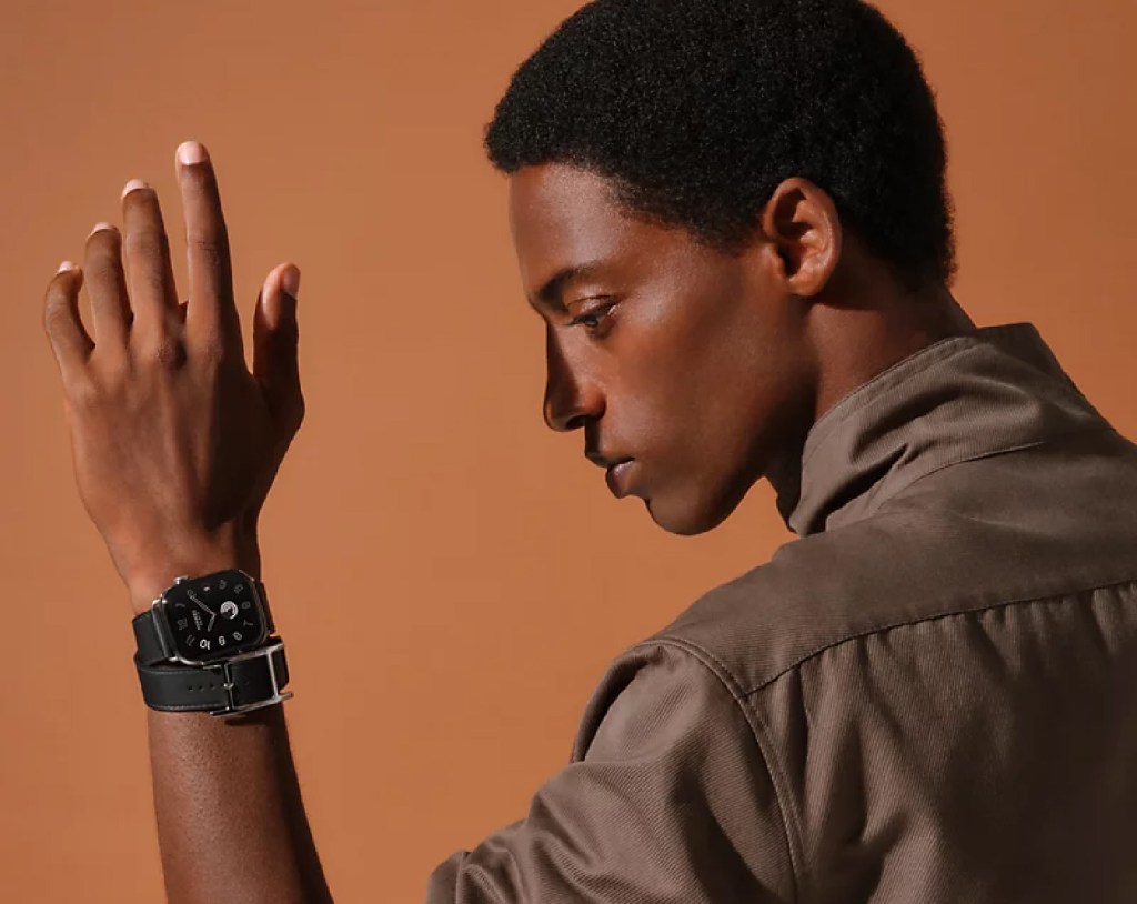 A close-up side profile of a person wearing a black smartwatch with a leather strap, raising their hand against a warm, neutral background.