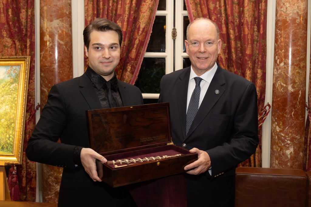 Two men, one in formal attire holding a wooden box, standing in a richly decorated room.