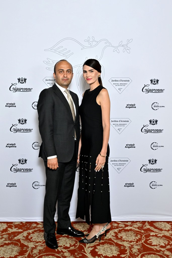A man in a formal suit stands next to a woman in an elegant black dress, posing in front of a branded backdrop for the Jardins d'Arménie Royal Brandy launch event.