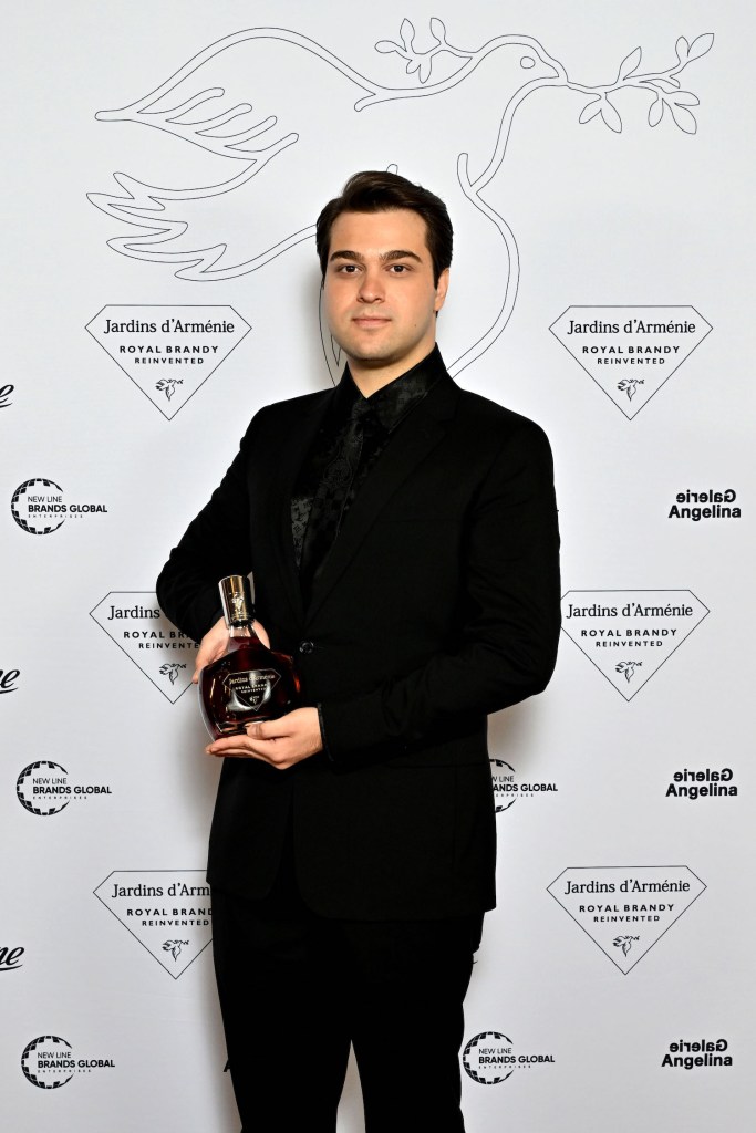 A young man in a black suit holds a bottle of Jardins d'Arménie Royal Brandy, standing against a backdrop featuring branding elements and a dove graphic.