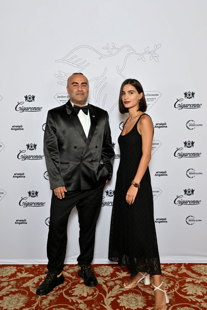 A man in a black tuxedo and a woman in a black dress pose together in front of a white backdrop featuring logos for Jardins d’Arménie, Galerie Angelina, and Cigaronne.