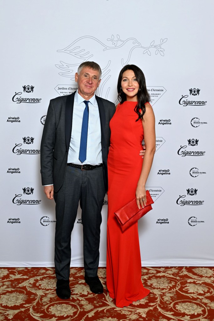 A man in a suit and a woman in a red dress pose together in front of a decorative background with branding for Jardins d'Arménie and Cigaronne.