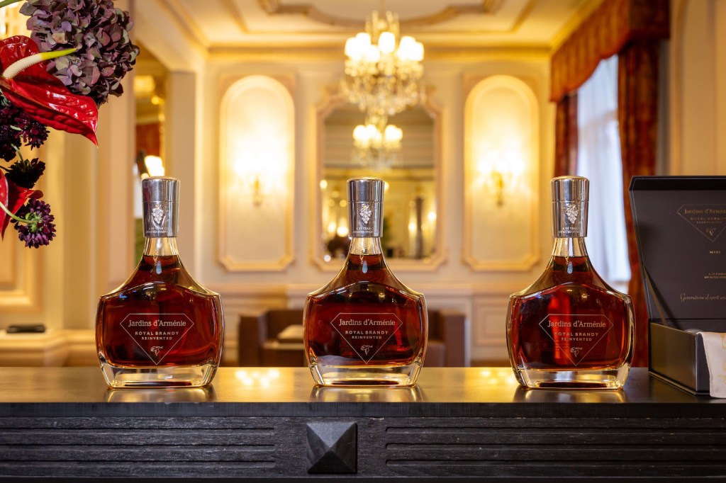 Three bottles of Jardins d'Armenie Royal Brandy displayed on a polished wooden surface, set against an elegantly decorated backdrop with chandeliers and soft lighting.