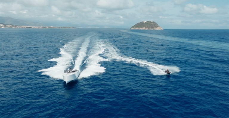 A yacht and a jet ski create white trails on the vibrant blue sea under a partly cloudy sky. An island is visible in the distance, evoking a sense of adventure.