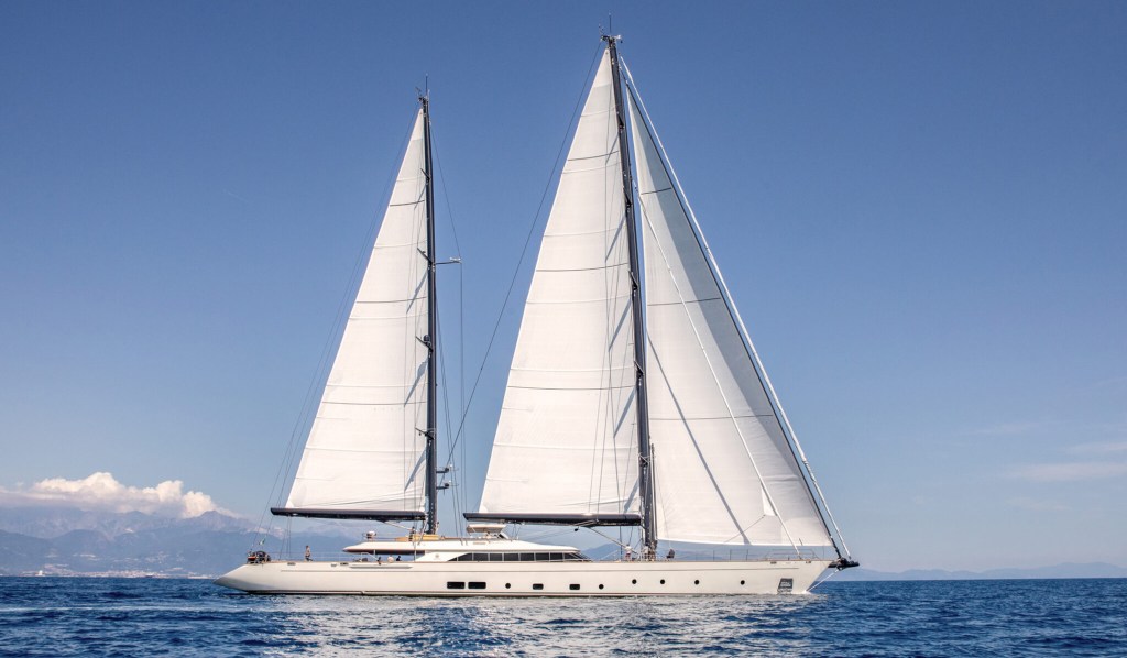 A large white sailing yacht glides on a calm, blue sea under a clear sky. The two tall sails are fully unfurled, and distant mountains are visible.