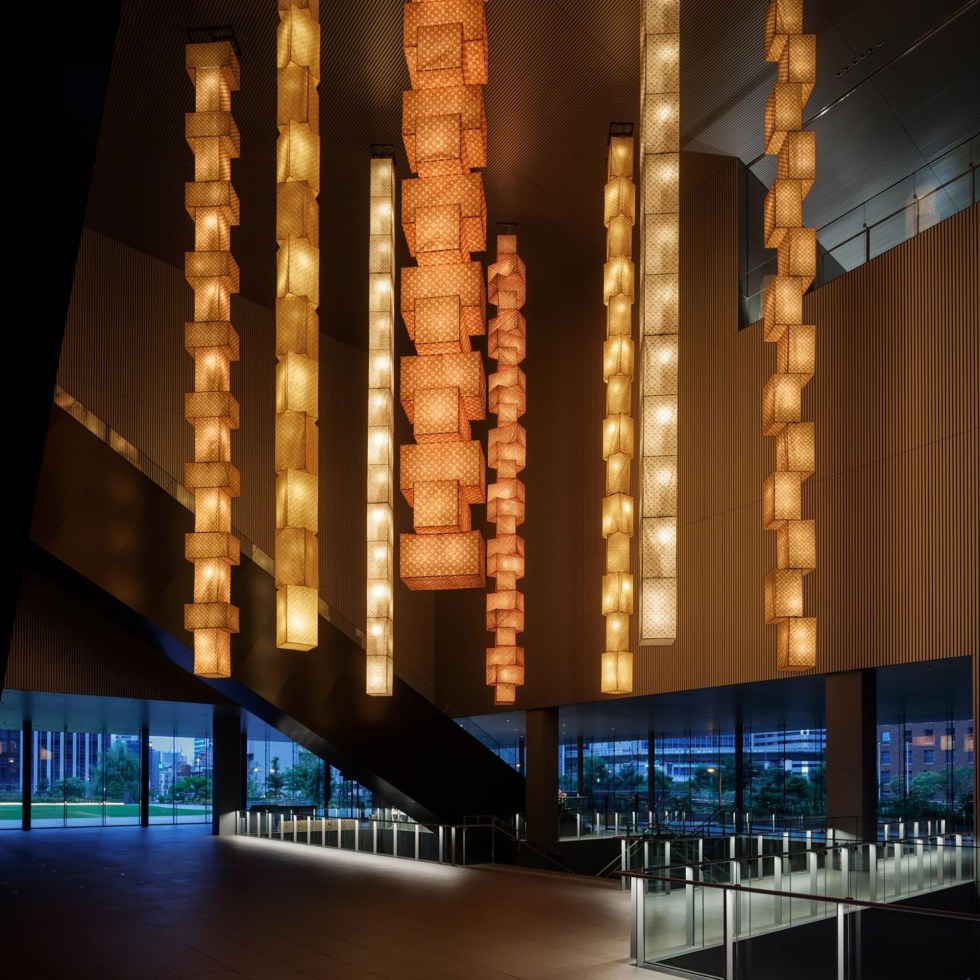 Illuminated pendant lights hang in a modern lobby, casting warm glows against sleek walls and large windows revealing the evening skyline.