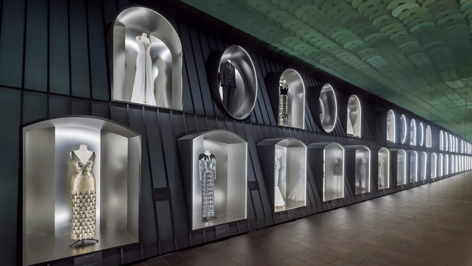 A long hallway displays fashion pieces in illuminated arched niches. Elegant dresses and garments are showcased against a sleek, modern backdrop, evoking sophistication.