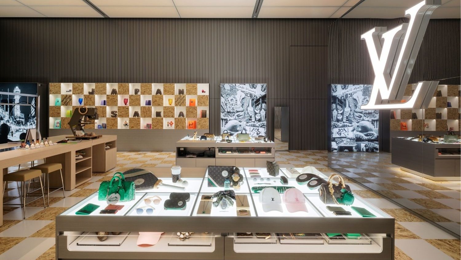 A luxurious retail interior featuring well-lit glass display tables with designer handbags and accessories. An illuminated "LV" logo hangs prominently, set against modern decor.
